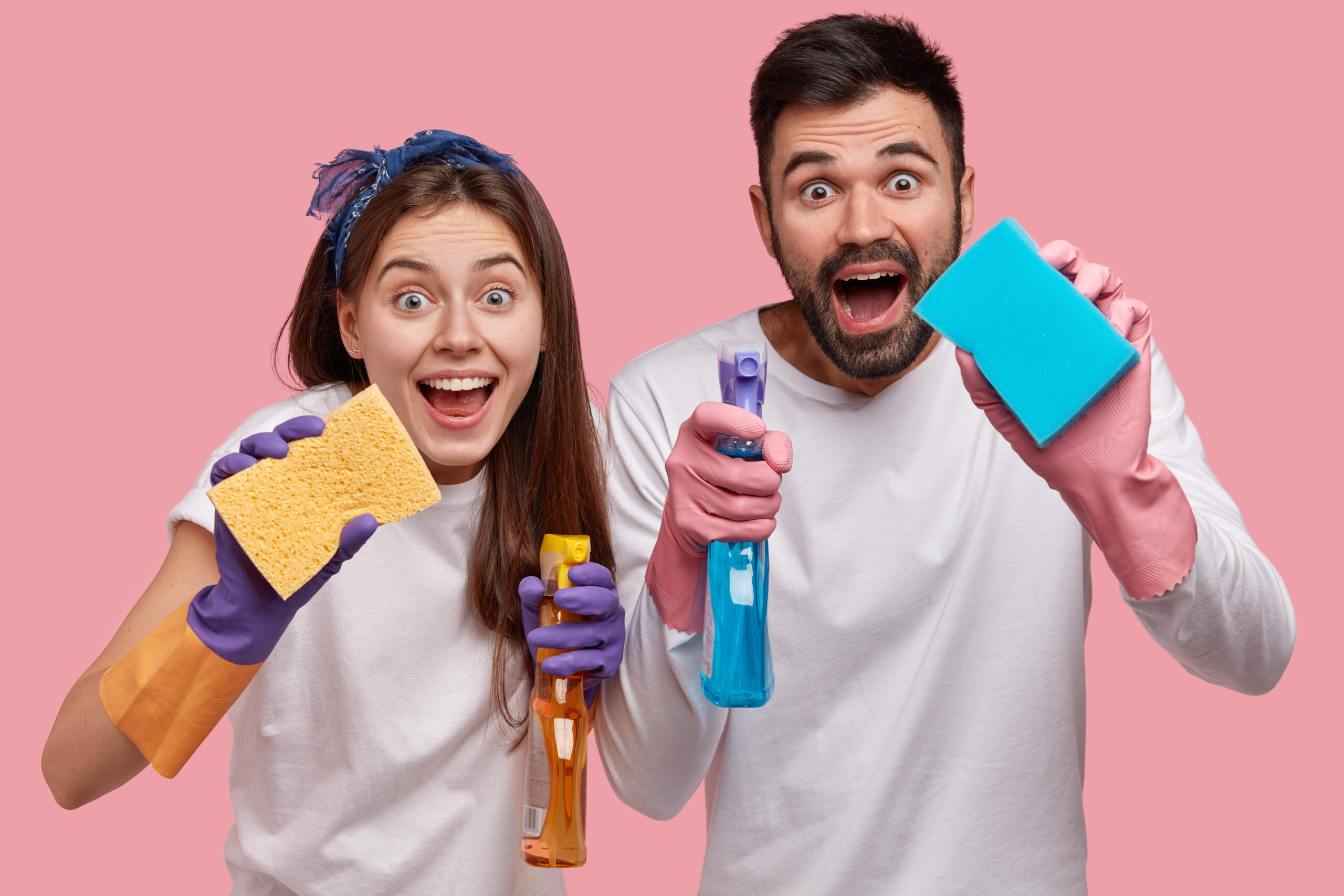 Positive young family couple have positive facial expressions, use chemical washing spray and sponge for cleaning window in room, have fun together while do household duties, isolated on pink wall.